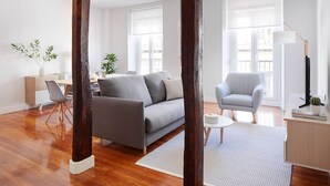 Ein modernes Wohnzimmer mit grauem Sofa, einem weißen Sessel und einem kleinen runden Tisch. Zwei Fenster mit weißen Jalousien, Holzboden und weiße Wand.