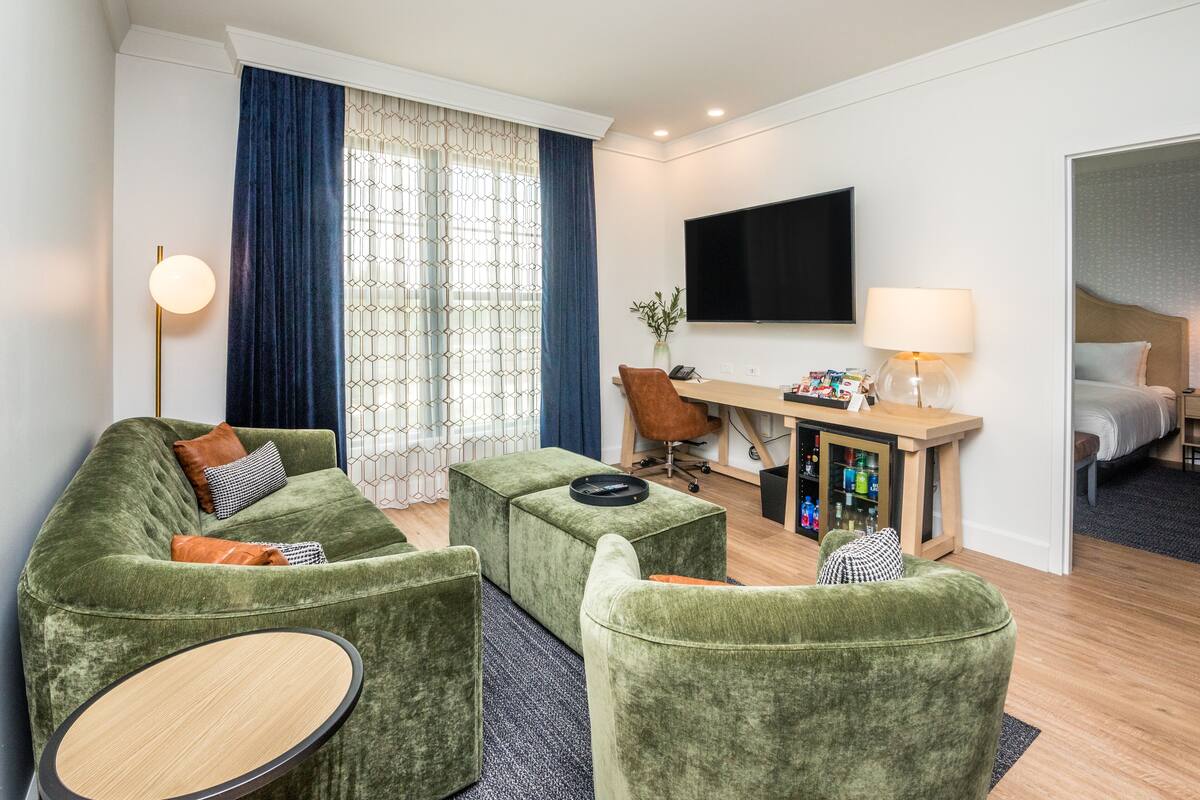 Una habitación de hotel moderna con un sofá verde, una mesa de centro de madera, un escritorio con una lámpara y un televisor de pantalla plana.