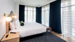 A modern hotel room with a large bed, a bedside table with a lamp, and a view of the city through patterned curtains.