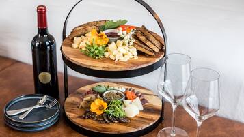 Un plateau de service en bois à deux niveaux, garni de différents fromages, de fruits et de pain, accompagné d’une bouteille de vin et de deux verres à vin, le tout posé sur une table.