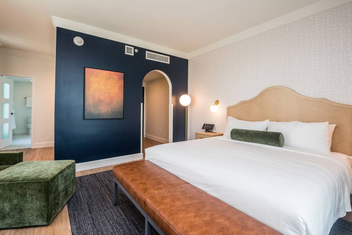 Una habitación de hotel con una cama grande, un banco, un escritorio y un cuadro en la pared.