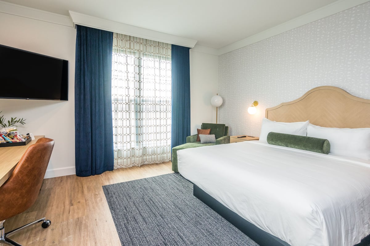 Habitación de hotel con cama, escritorio, silla, televisión y ventana con cortinas.