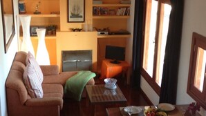 A cozy living room with a sofa, a coffee table, and a fireplace. There are shelves with books and decorative items, and a window with curtains.