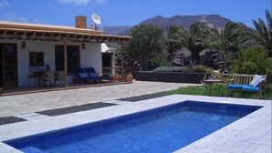 A pool area with a clear view of mountains in the background, a patio with outdoor furniture, and a building with a satellite dish.
