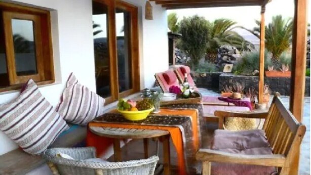 A covered outdoor seating area with wicker furniture, a table with a bowl of fruit, and a view of a garden with palm trees.