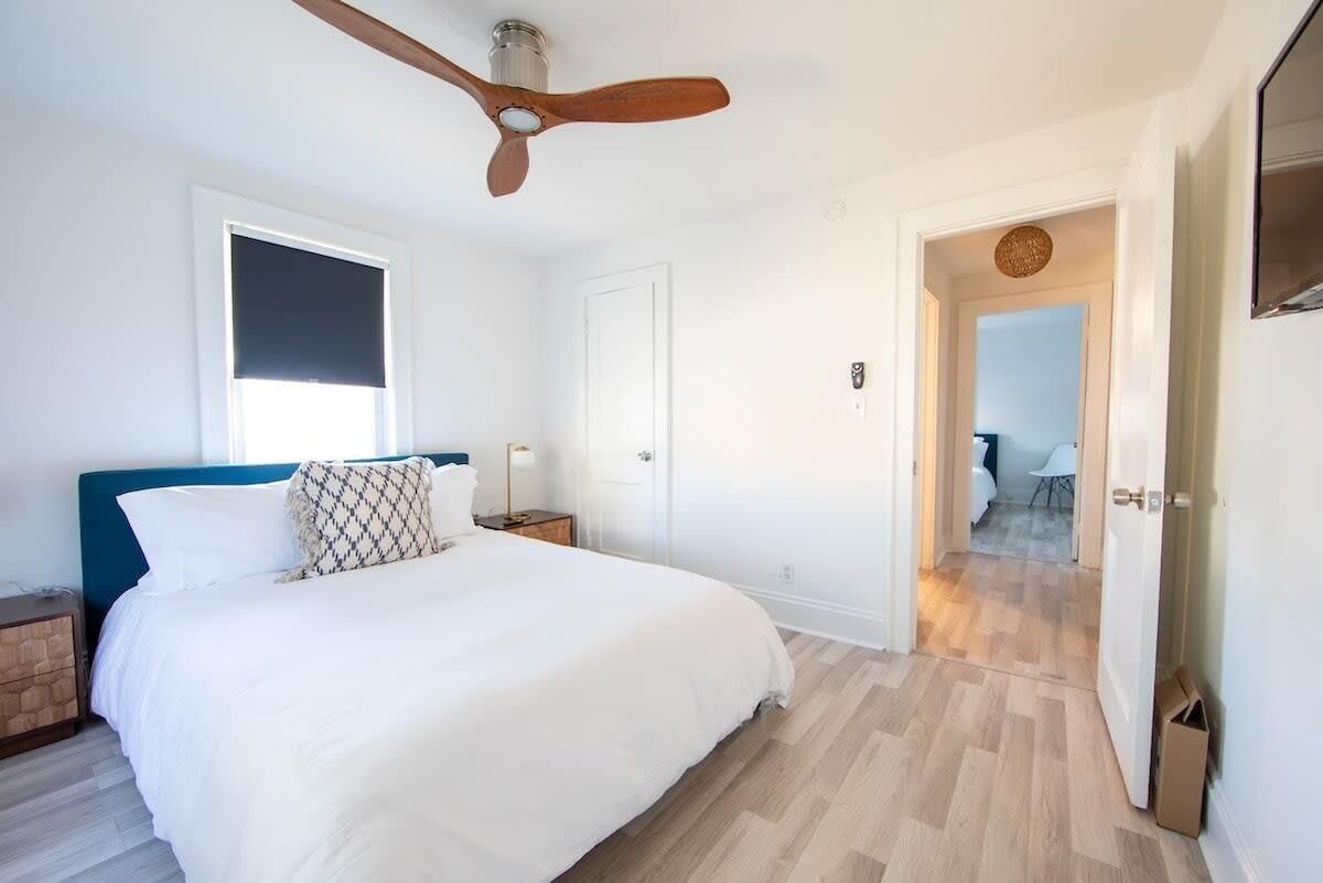 A bedroom with a bed, a ceiling fan, a window with blinds, and a door leading to another room.