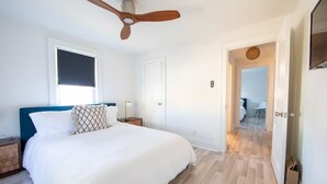 Quarto com cama, ventoinha no teto, janela com persianas e porta que dá para outro cômodo.