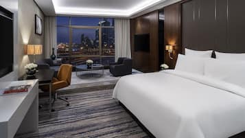 A hotel room with a large bed, a desk, a chair, a coffee table, and a view of the cityscape at night.