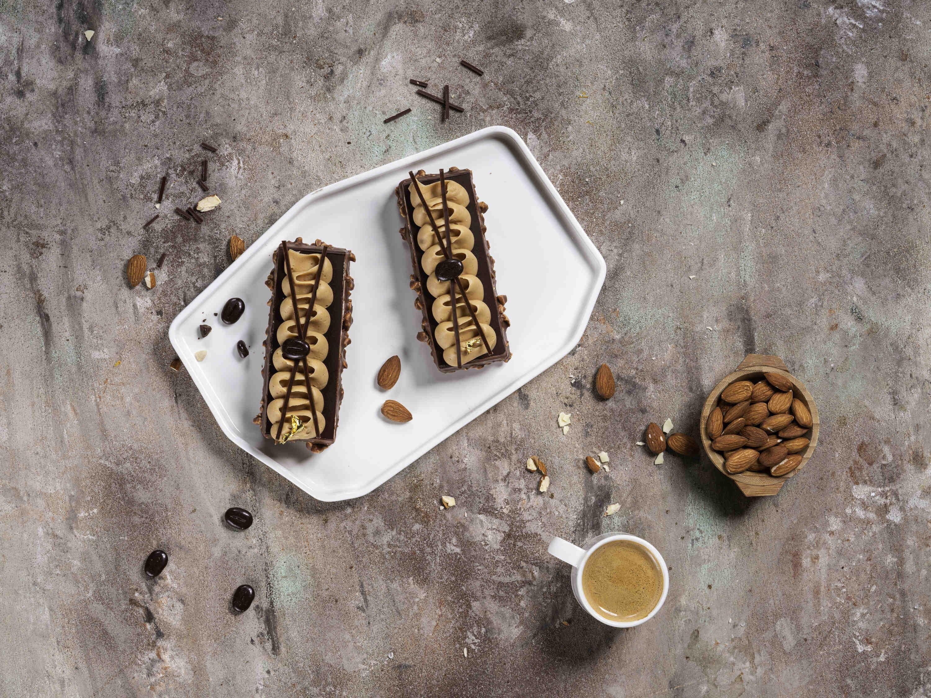 Deux desserts rectangulaires au chocolat sont présentés sur une assiette blanche, des amandes sont éparpillées autour, et une tasse de café repose sur une surface texturée.