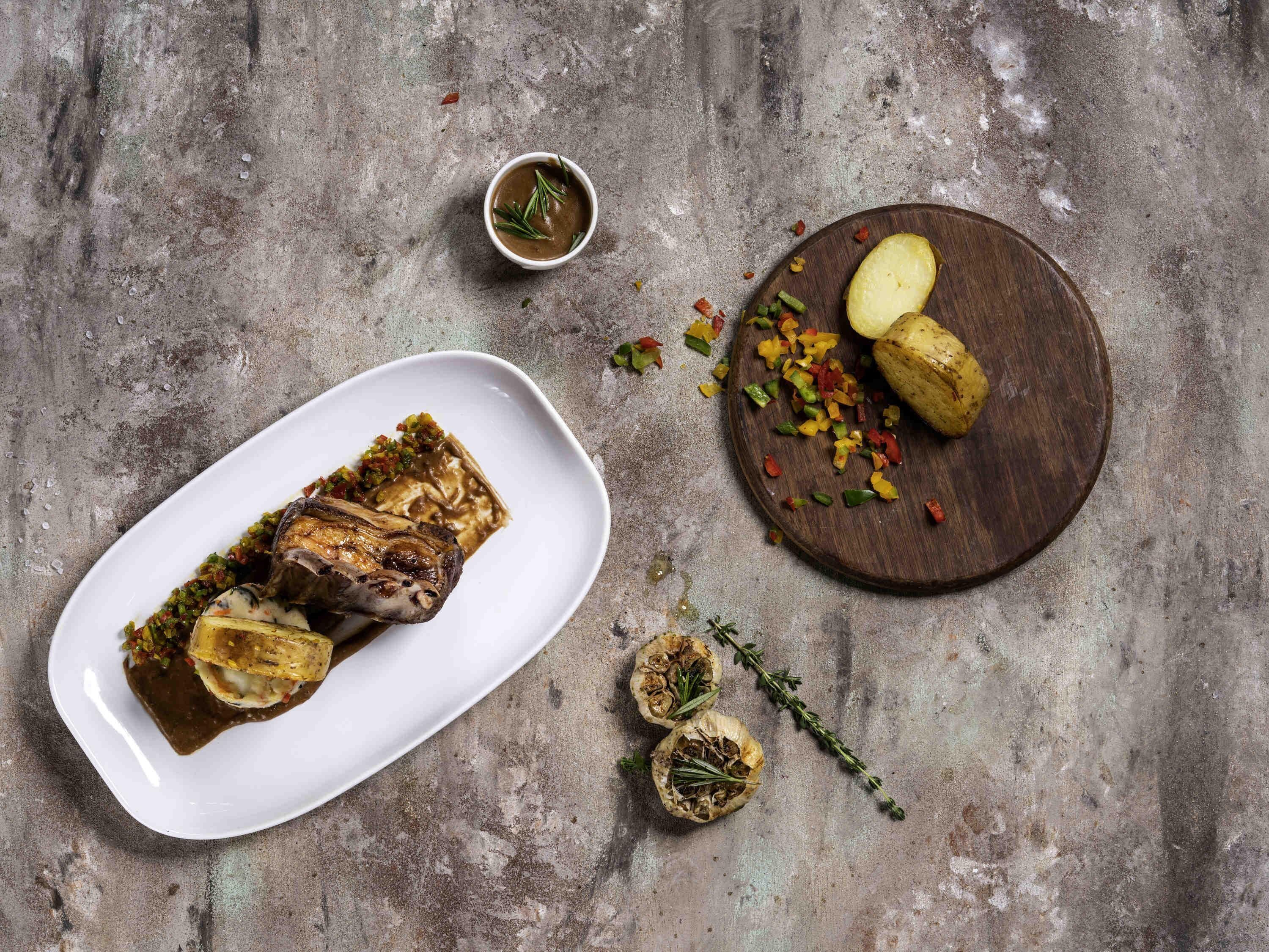 A meal with a white plate containing a roasted meat dish, a wooden board with sliced potatoes and diced vegetables, and a small bowl of sauce.