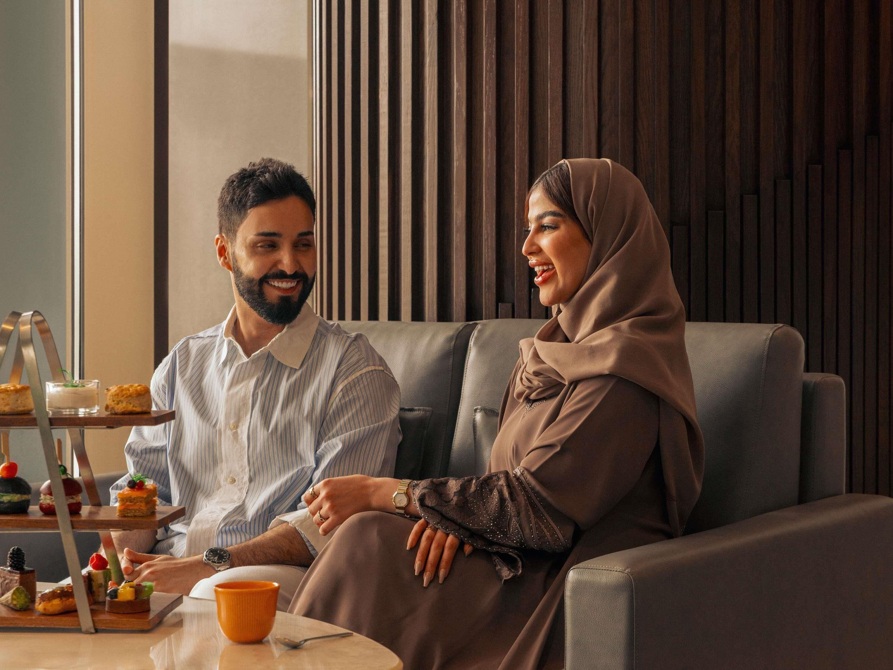 A couple seated on a sofa with a tray of desserts and a cup on a table.