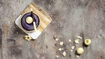 Una tabla de madera con un tazón morado y una cuchara de madera, rodeada de nueces picadas y una flor.