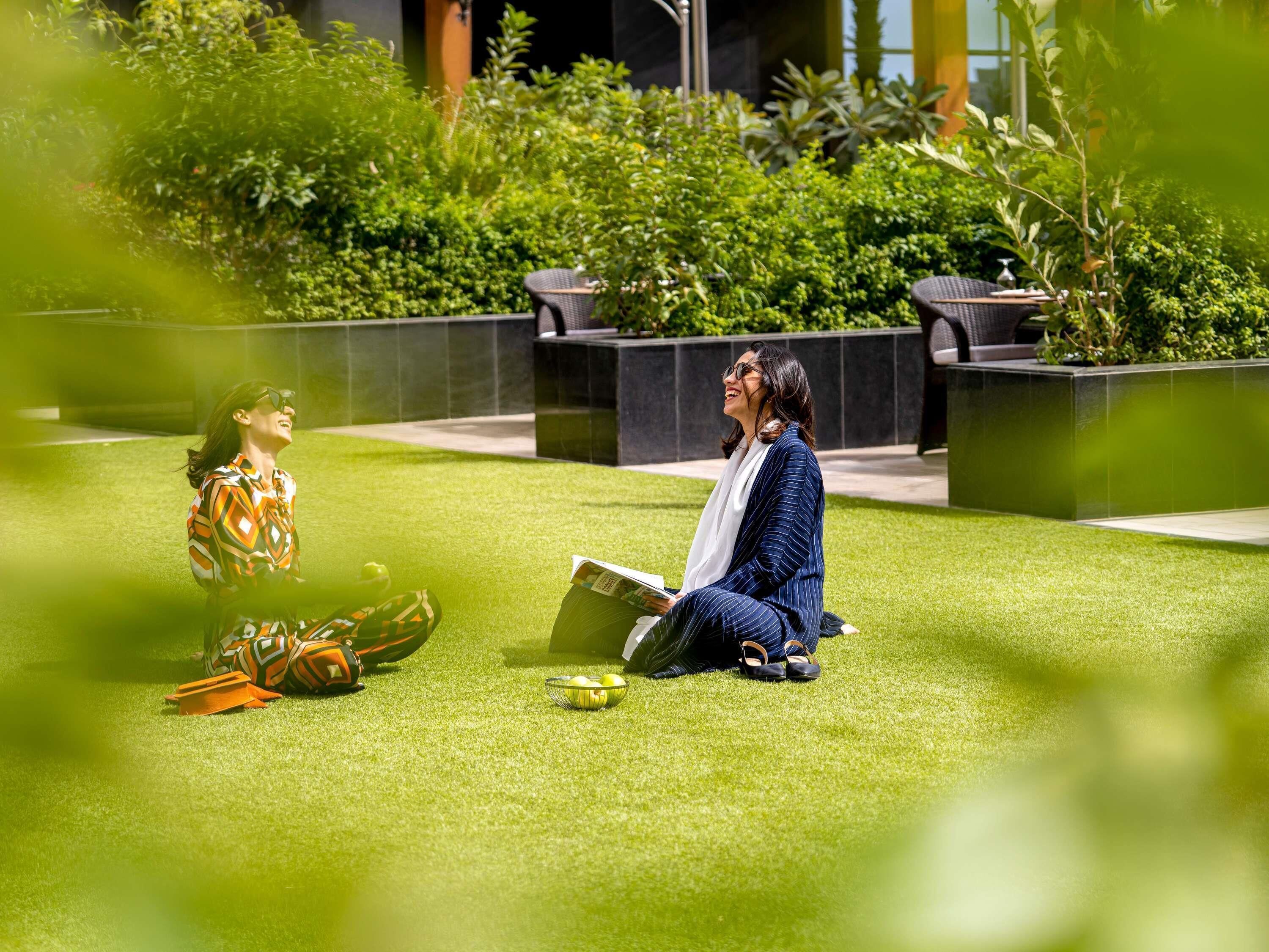 Two people sitting on artificial grass outdoors with a building and greenery in the background.