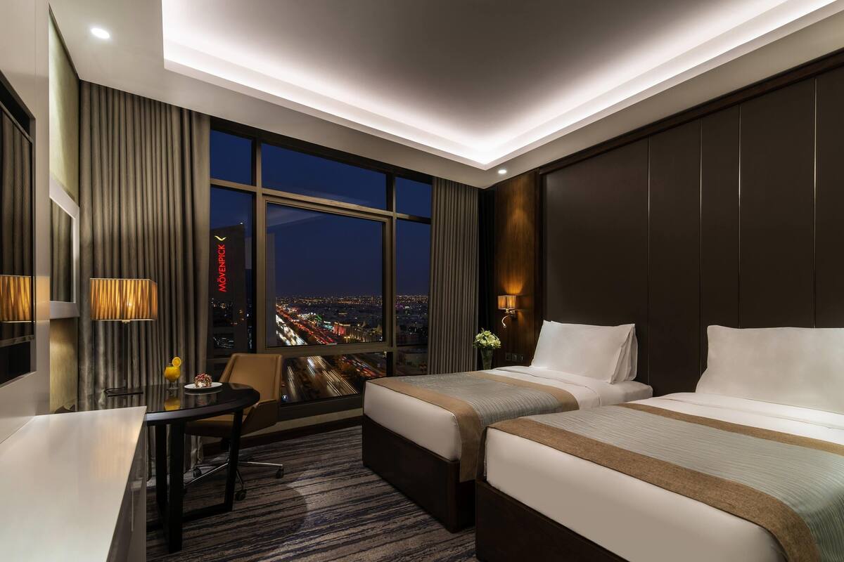 A hotel room with two beds, a desk, a chair, a large window with a city view, and a modern ceiling design.