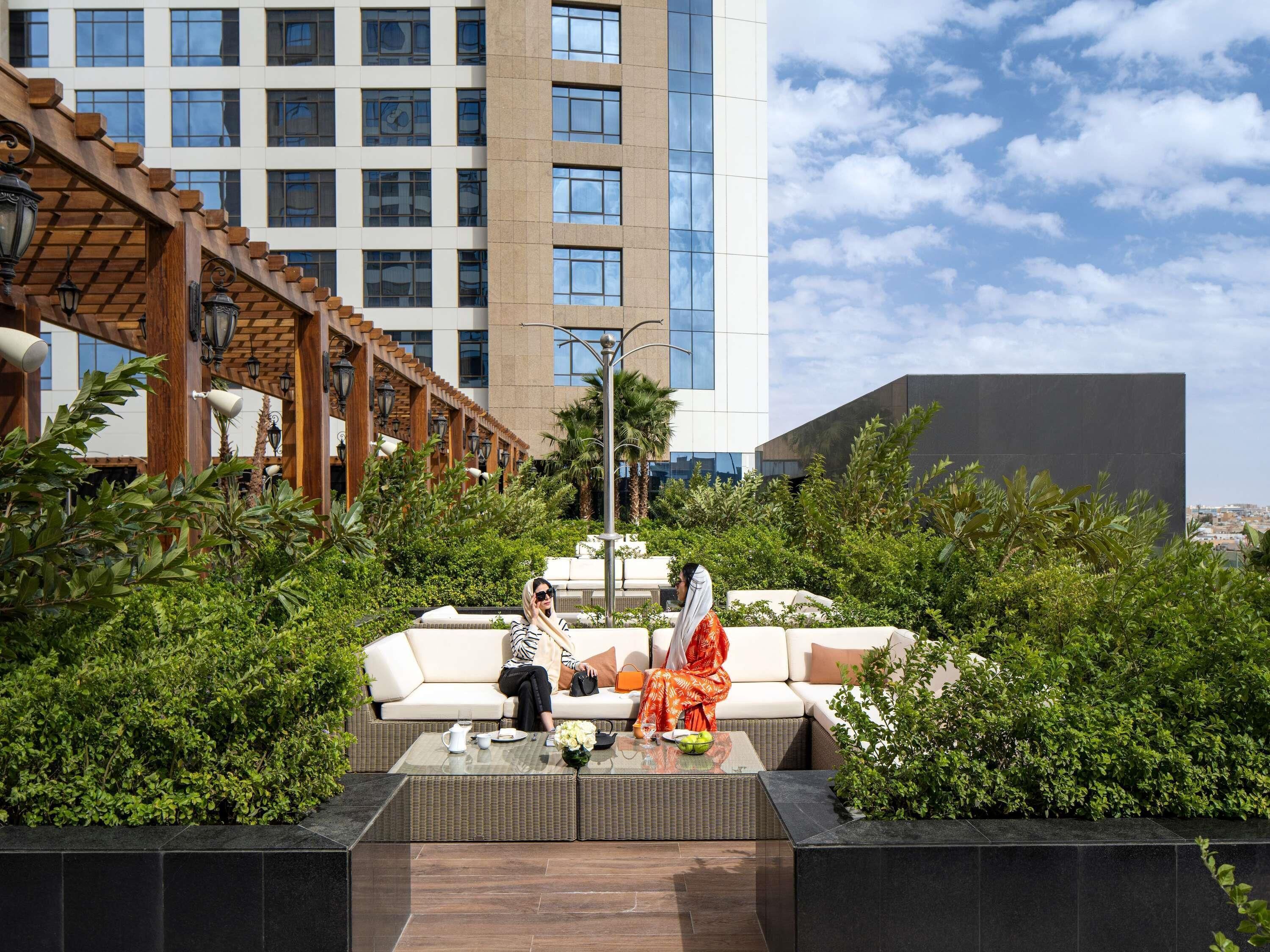 Two people sitting on a white outdoor sofa with a glass table in front, surrounded by greenery and a modern building in the background.