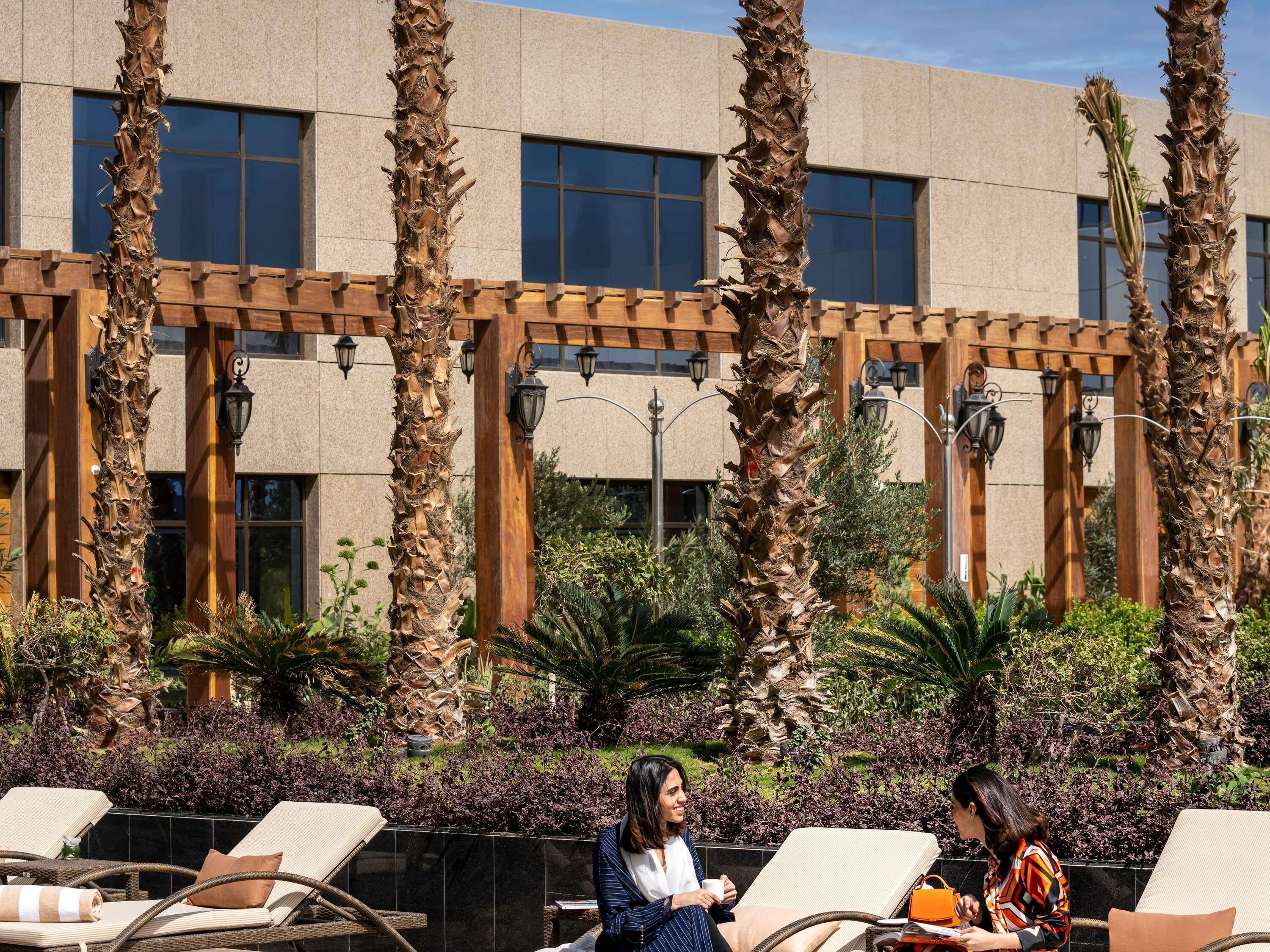 A pool area with lounge chairs, palm trees, and a building with large windows and outdoor lighting.