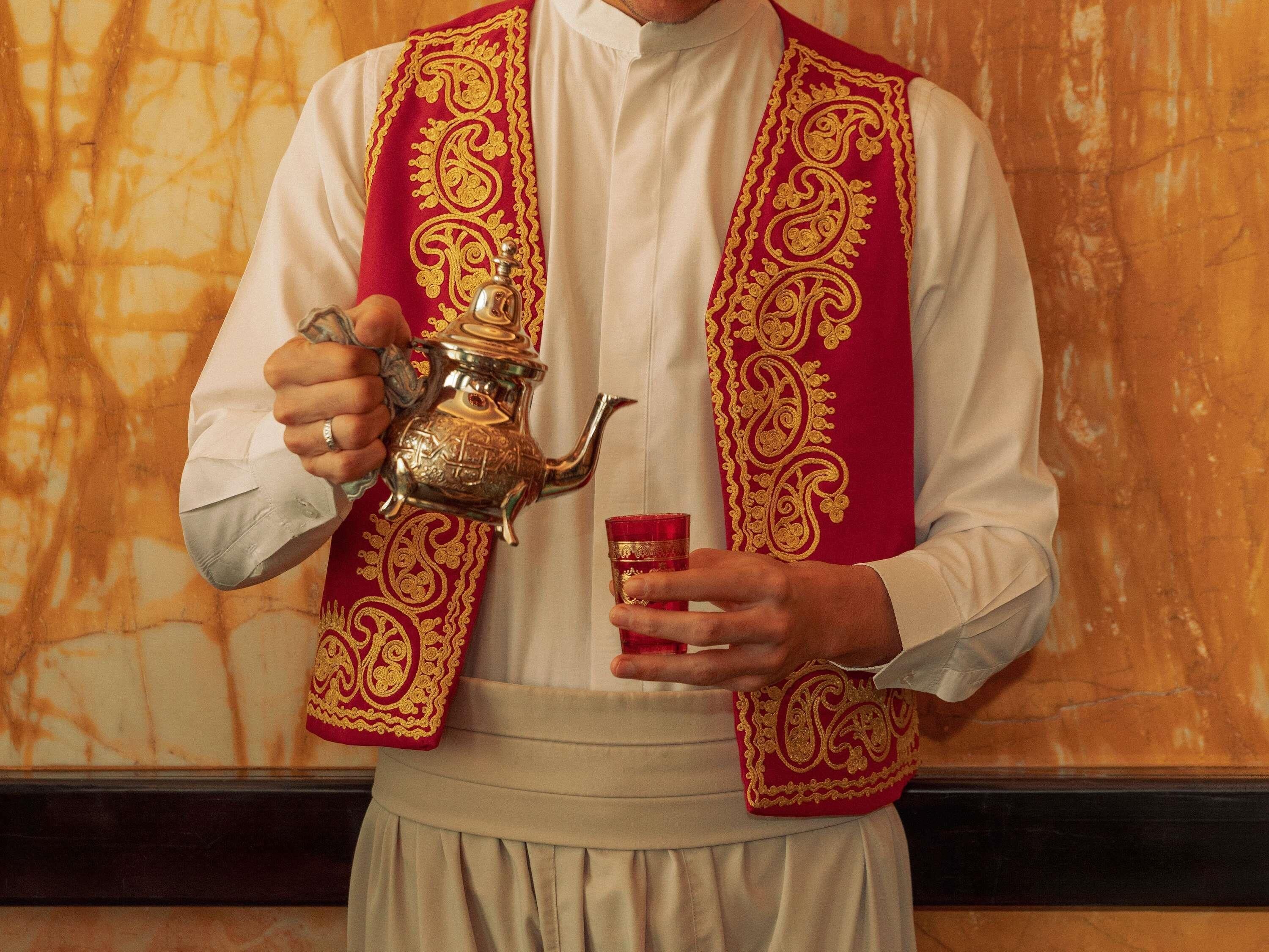 A person in traditional attire holding a teapot and a cup, with a patterned garment and a white shirt.