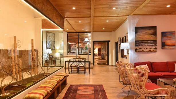 A spacious living area with a wooden ceiling, a dining table with chairs, a red sofa, wicker chairs, and a bench with cushions.