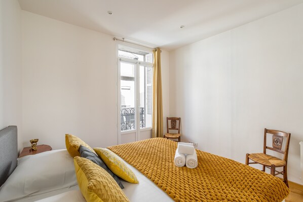 Um quarto com cama, janela com cortinas, uma cadeira e uma mesinha.