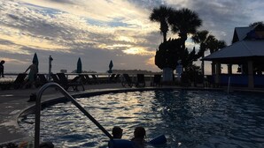 Ein Swimmingpool mit Liegestühlen und Sonnenschirmen, eine Palme und ein Gebäude mit überdachtem Bereich.