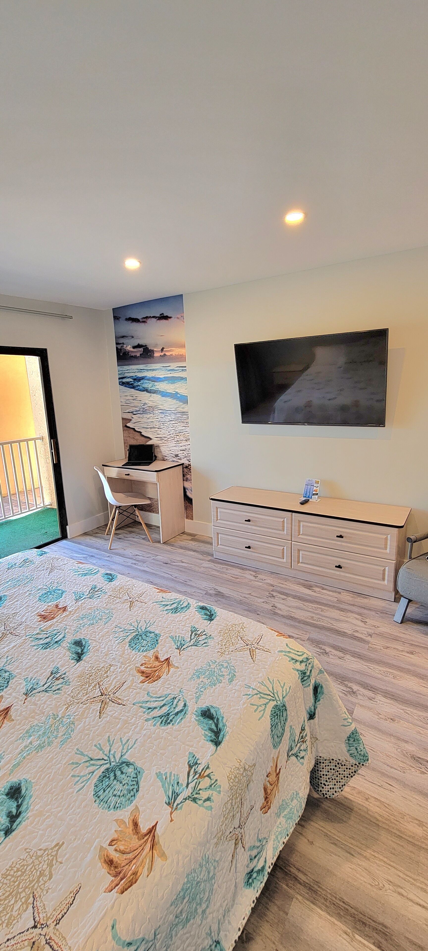 Un dormitorio con cama, escritorio, cómoda y un televisor grande montado en la pared.