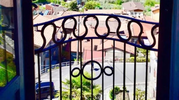 Een balkon met een metalen hek dat uitkijkt over een bergachtig landschap met een meer en gebouwen.