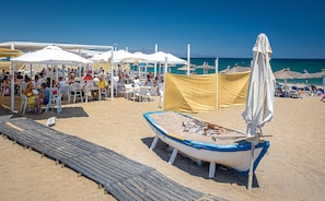 A beach with a row of white umbrellas, a wooden walkway, and a small boat with a blue and white hull.