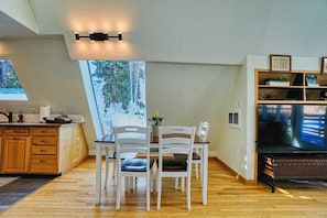 A dining area with a wooden table and white chairs, a kitchen with wooden cabinets, and a TV on a stand. - Creekside Mountain Chalet with Hot Tub! Extended Stay (Black Hawk)