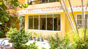 A yellow building with large windows, surrounded by lush greenery and potted plants.