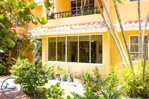 A yellow building with large windows, surrounded by lush greenery and potted plants.