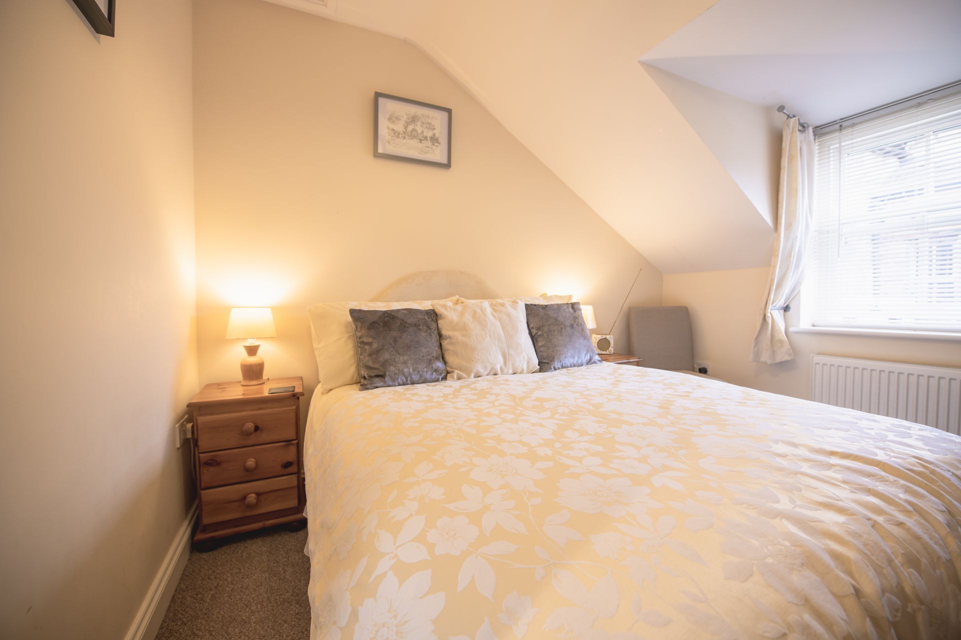 Ein Schlafzimmer mit einem großen Bett, einem Nachttisch, einer Lampe, einem gerahmten Bild an der Wand und einem Fenster mit Vorhängen.