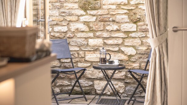 A stone wall, two chairs, a table with a coffee pot and cup, and a window with curtains.