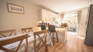 A compact kitchen with wooden cabinets, a dining area with a table and chairs, and a view of a stone wall through an open door.