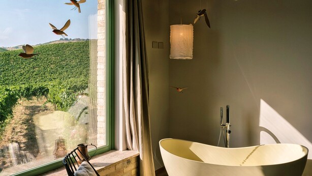 A modern bathroom with a freestanding bathtub, a window with a view of a vineyard, and a hanging light fixture. - Cocci Grifoni - Panoramic Wine Resort (Offida)