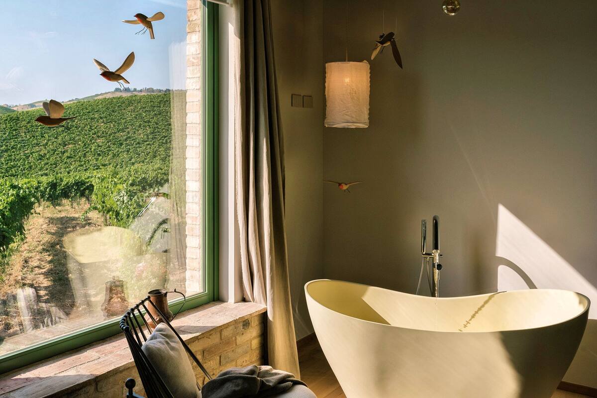 A modern bathroom with a freestanding bathtub, a window with a view of a vineyard, and a hanging light fixture.