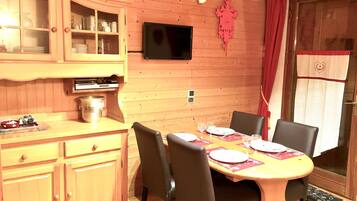 Een houten eetgedeelte met een tafel gedekt voor vier personen, stoelen, een aan de muur bevestigde televisie en een kast met glazen deuren.
