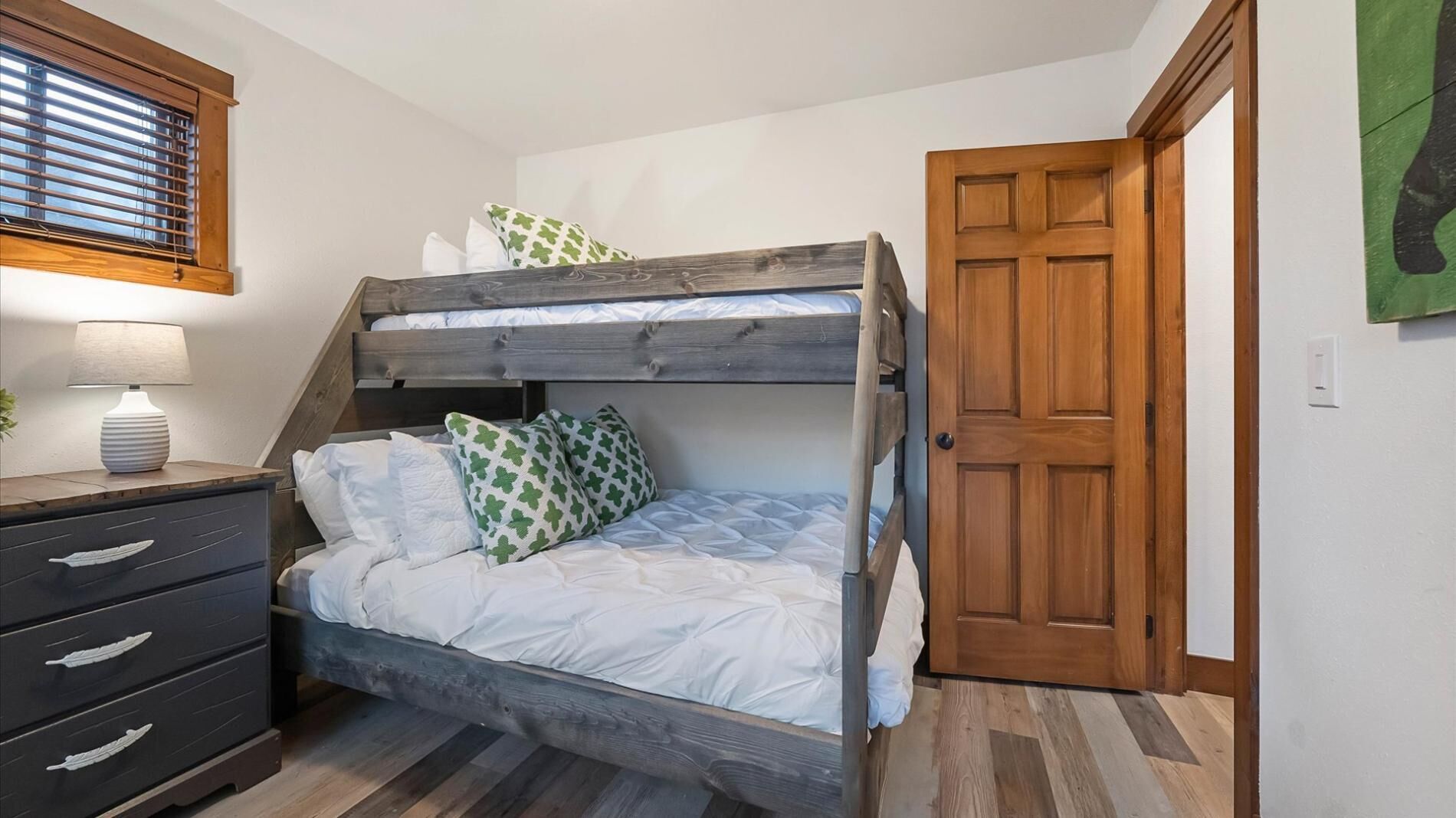 Un lit superposé avec une literie blanche et des oreillers à motifs verts, une lampe de chevet, une commode en bois avec des tiroirs et une porte en bois.