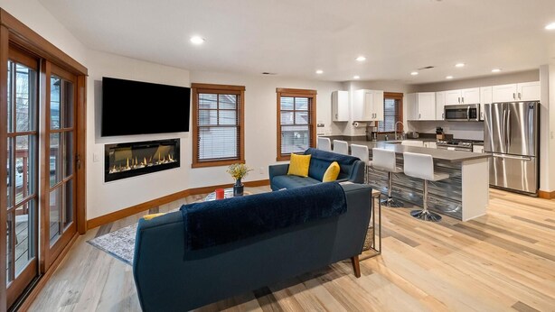 A modern kitchen with stainless steel appliances, a dining area with a bar, and a living room with a blue sofa and fireplace.