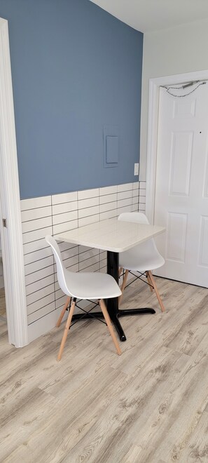 A dining area with a white table and chairs, a blue wall, and a white door. - Satellite Motel - Happy Daze 107 (Treasure Island)