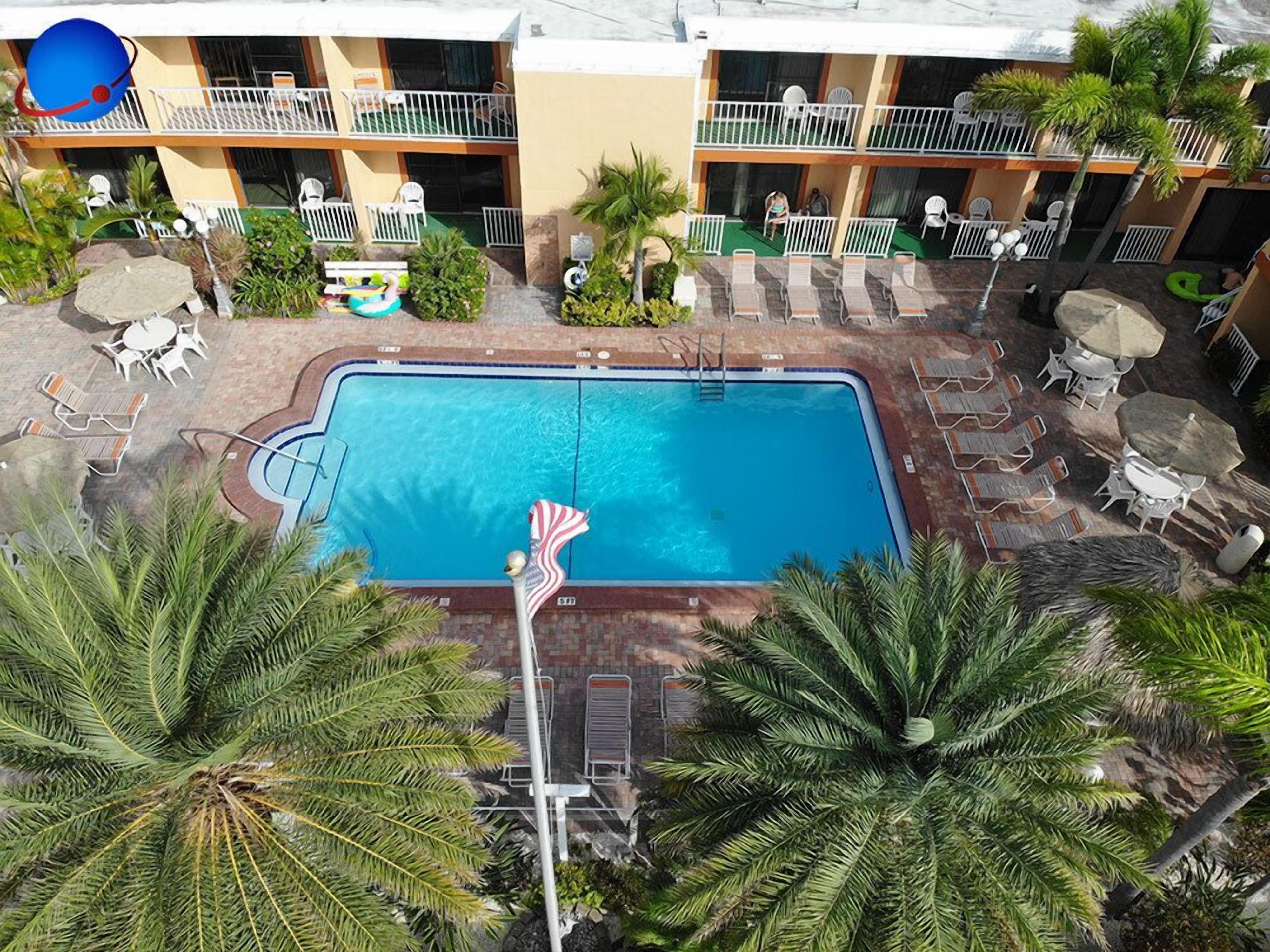 Um hotel com piscina, rodeado por espreguiçadeiras e guarda-sóis, palmeiras e um edifício de vários andares ao fundo.