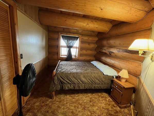 A cozy log cabin bedroom with a bed, a fan, a lamp, and a window with curtains.