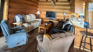 Interior de una cabaña de troncos con techo de madera, un sofá, un sillón, una mesa de centro, un televisor y una pequeña área de cocina.