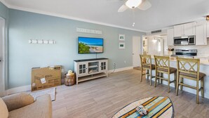 Un salon avec un téléviseur, un coin repas avec des chaises, une cuisine équipée de placards et d’un four à micro-ondes, et un ventilateur de plafond.
