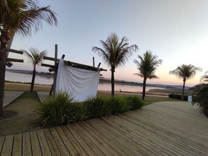 A wooden deck with a white cloth draped over a wooden structure, palm trees, and a view of a river. - Country house with solar-heated swimming pool and barbecue area. (Itaí)