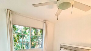 Une chambre à coucher avec un lit, un ventilateur de plafond, une fenêtre avec des rideaux et une vue sur la verdure à l’extérieur.