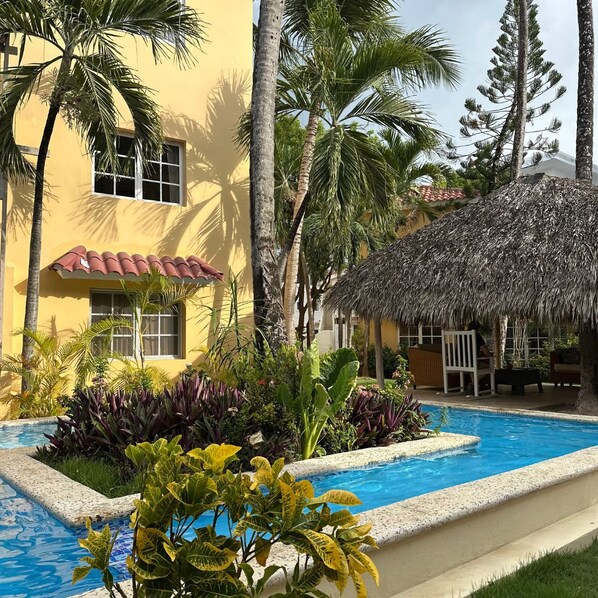 A pool area with a thatched roof structure, surrounded by tropical plants and palm trees.
