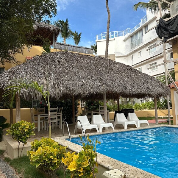 A swimming pool with a thatched umbrella, white lounge chairs, and a building in the background.