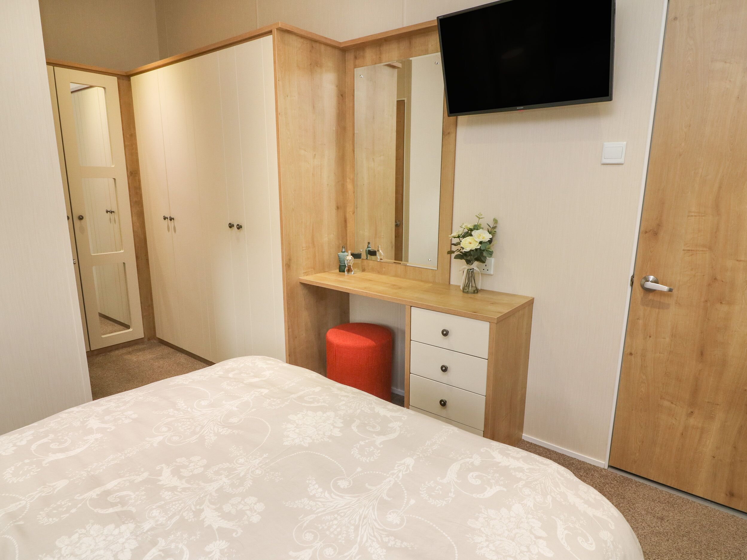 Un dormitorio con una cama, un aparador de madera, un espejo, un televisor y un pequeño florero con flores.