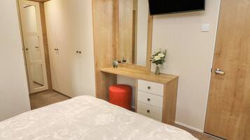 Un dormitorio con una cama, un aparador de madera, un espejo, un televisor y un pequeño florero con flores.