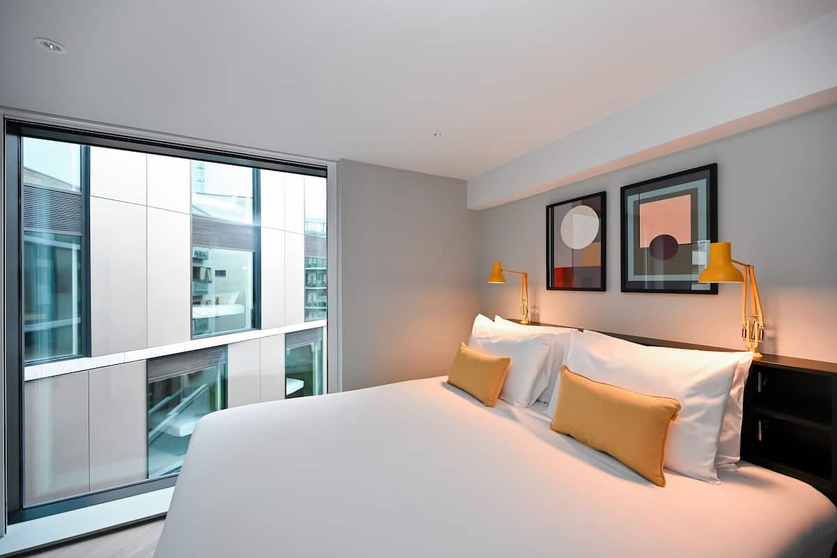 Ein modernes Hotelzimmer mit einem großen Bett, zwei Nachttischlampen und Blick auf die Stadt durch raumhohe Fenster.